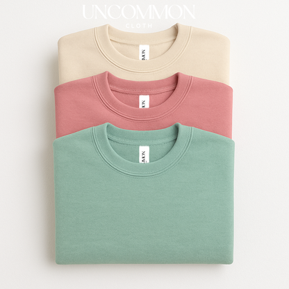 Three folded sweatshirts in beige, pink, and green on a white background with 'UNCOMMON cloth co' branding.